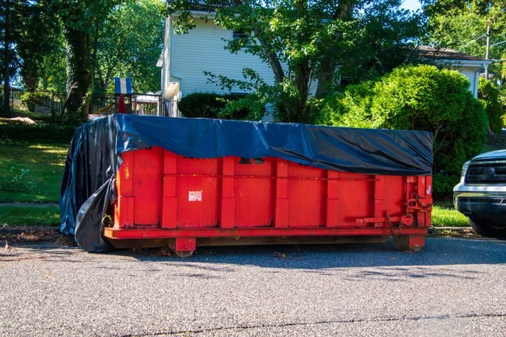 A large red dumpster, provided by a Plano, TX dumpster rental service, sits on a residential street, partially covered with a black tarp. Trees and a house are visible in the background.