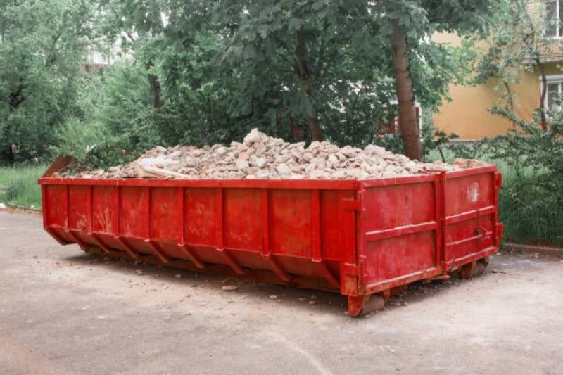 A large red dumpster filled with debris is positioned on a street surrounded by greenery.
