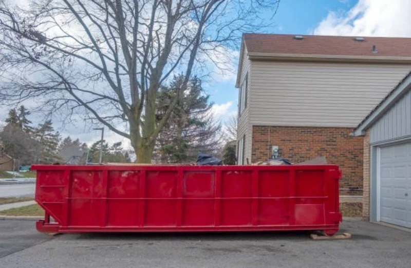 A red dumpster parked next to a house, with a tree and cloudy sky in the background.