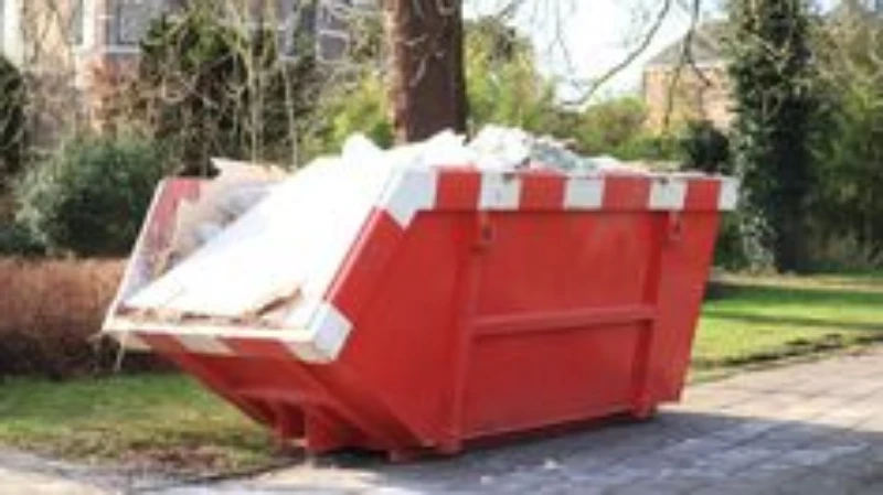 A red dumpster filled with construction debris, placed on a paved pathway next to greenery.