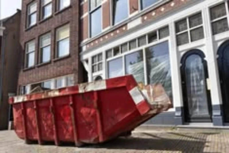 A red dumpster tipped over on its side in front of a building with large windows.