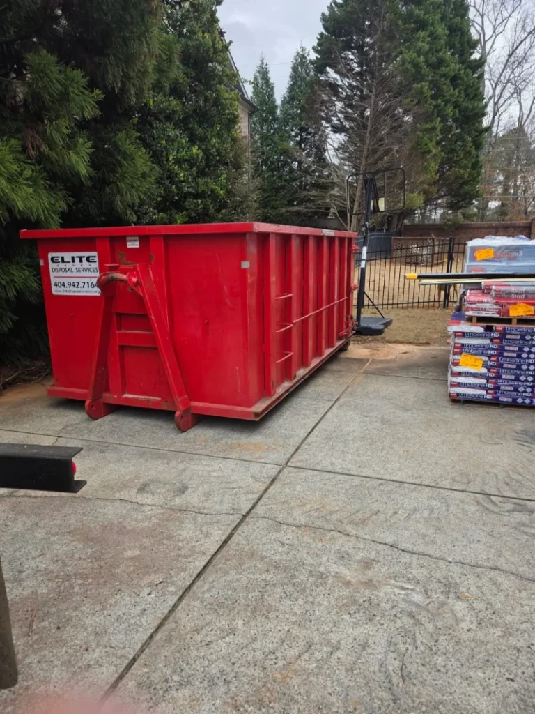 A large red dumpster labeled "ELITE" is parked on a concrete surface, with trees and a fence in the background.