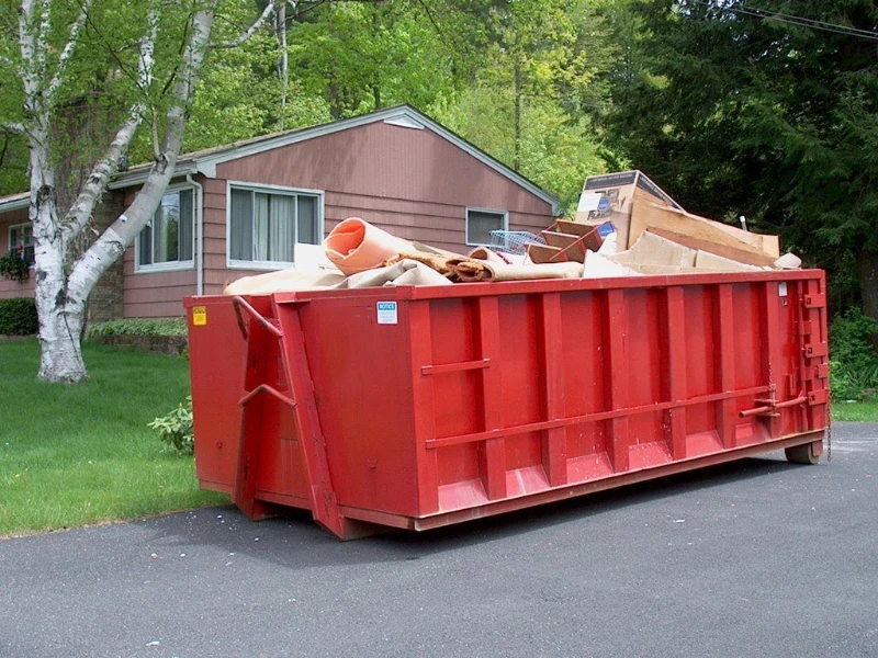 A large red dumpster filled with various types of debris and materials, placed on a paved surface beside a house surrounded by trees.