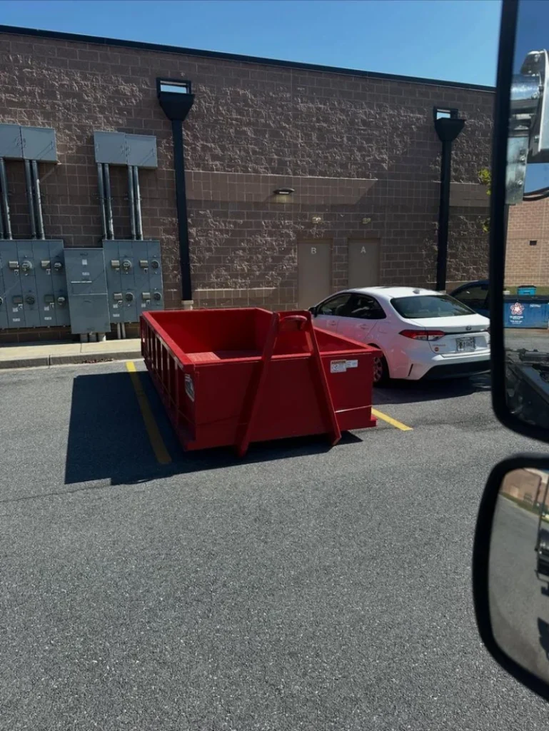 A red dumpster parked in a lot next to a white car, with utility boxes visible on the wall behind.