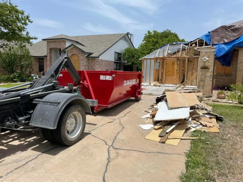 Red trash trailer beside yard debris, with construction materials and houses in background.