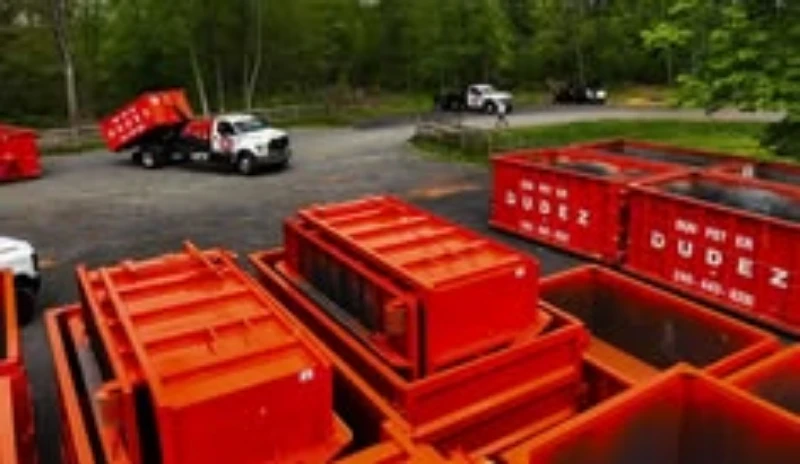 A landscape featuring several red dumpsters and a truck backing up to one of the dumpsters in a wooded area.