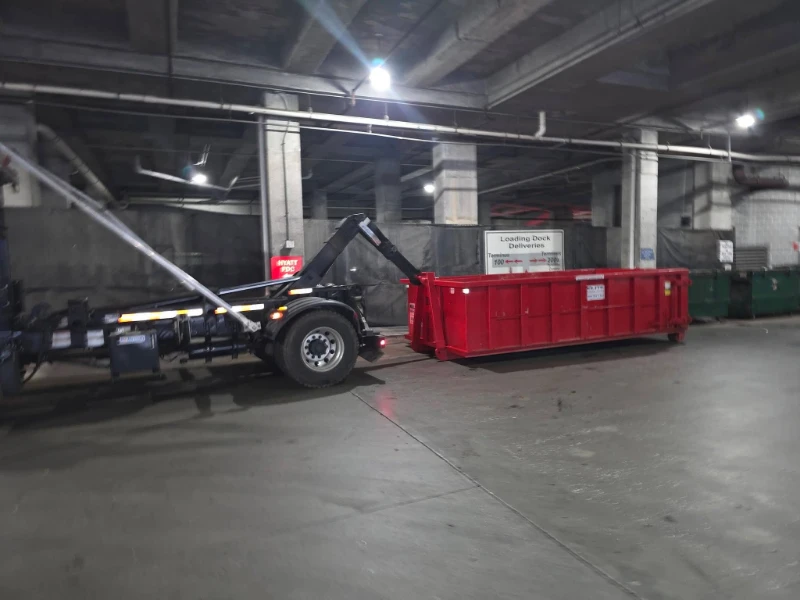 A truck is unloading a large red dumpster in an underground loading dock area with concrete pillars and overhead lighting.