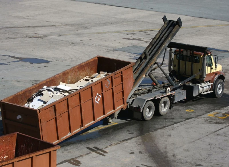 A truck is unloading a large metal dumpster filled with construction debris onto a paved lot.