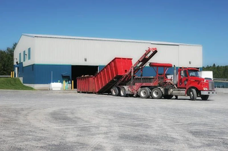 The image simply shows a red roll-off dumpster truck loading or unloading a large red container in front of a blue and white industrial warehouse. If you need any other details about what's pictured, feel free to ask!