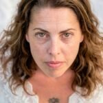Close-up portrait of a woman with a tattoo wearing a white top, looking directly at the camera.
