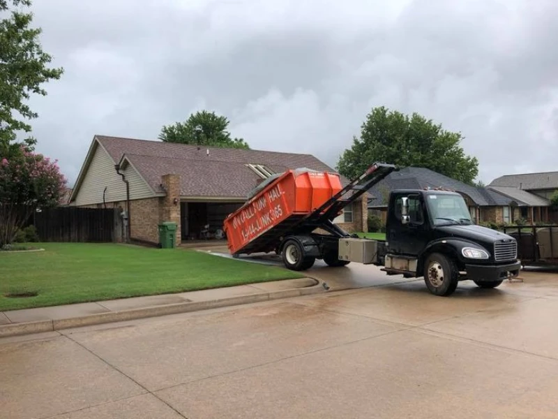 Black roll-off dumpster truck with an orange waste container parked on a wet residential street under a cloudy sky; quiet neighborhood scene.