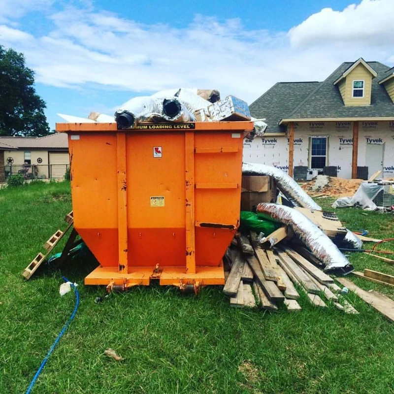 Orange construction dumpster overflowing with insulation and debris sits on a lawn beside a house under renovation beneath a bright blue sky.