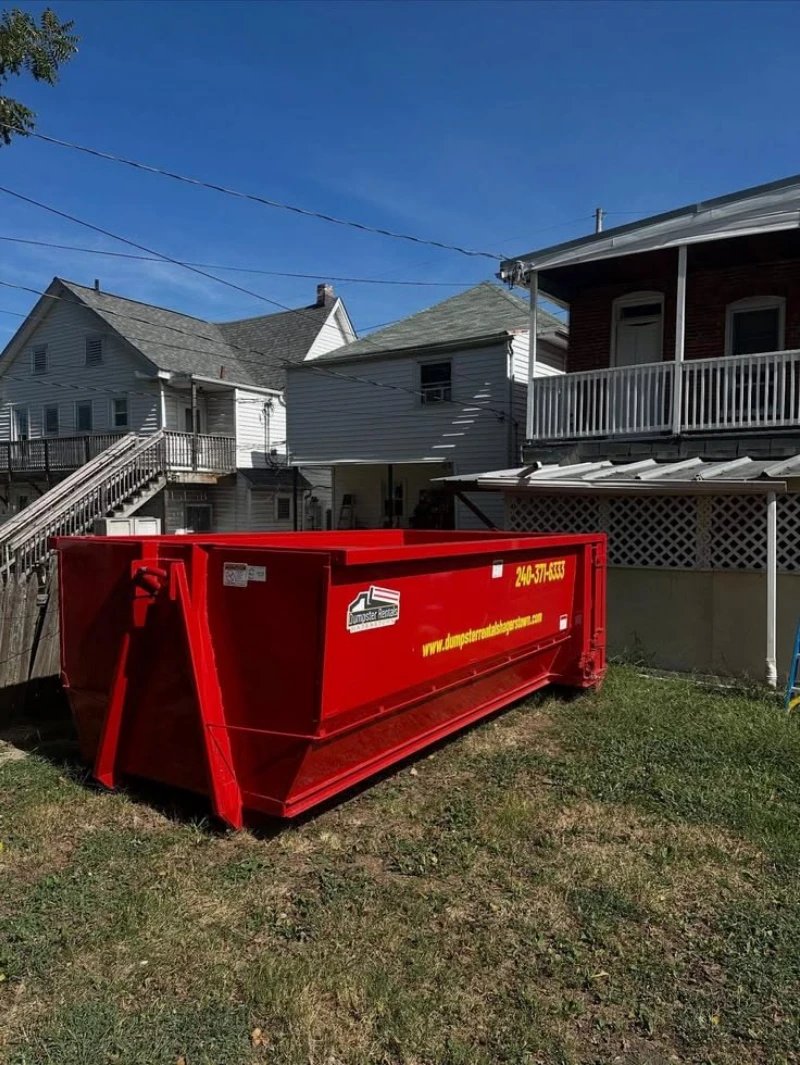 Bright red construction dumpster sits on a grassy lot between older gray homes beneath a clear blue sky, creating a sunny neighborhood scene.