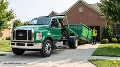 Green garbage truck with a raised cab and attached dumpster parked in a suburban driveway, with a worker nearby on a sunny day.