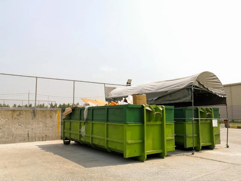Green industrial dumpsters filled with debris sit beneath a gray tarp in a fenced concrete yard, creating a utilitarian outdoor scene.