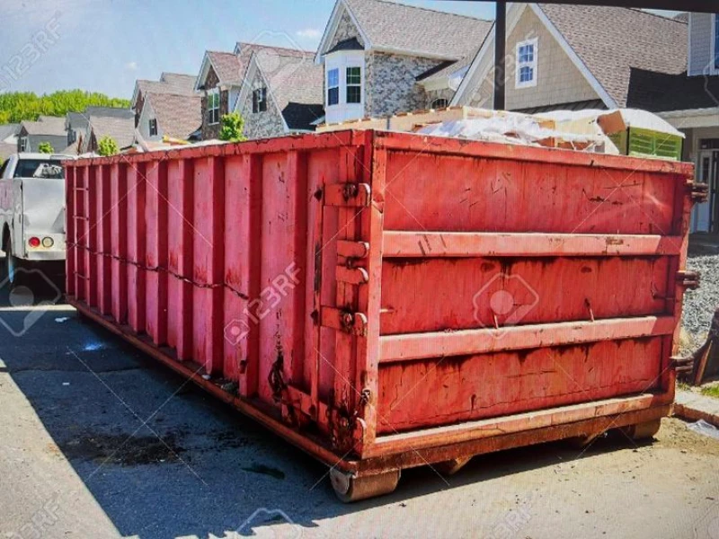 Large red construction dumpster filled with debris sits in a sunny parking area, with suburban townhouses and green trees behind it.