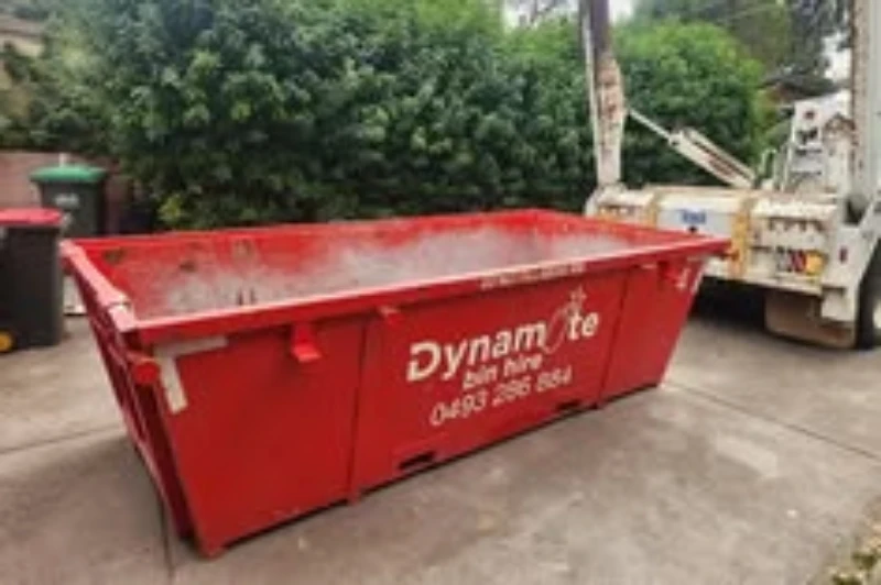 Bright red Dynamite skip bin sits in a driveway beside a garbage truck, with green trees and wheelie bins behind it; everyday cleanup scene.