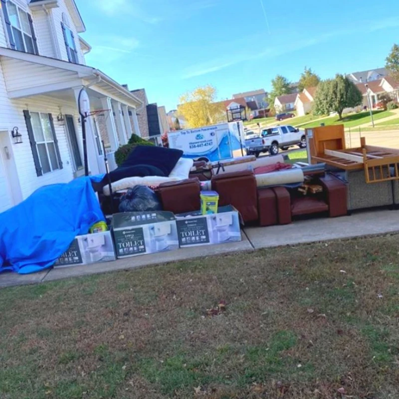 Curbside donation pickup with sofas, chairs, boxed toilets, and covered items outside a white home on a sunny suburban street.