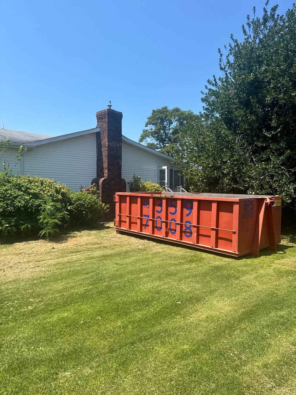 Bright orange construction dumpster with blue graffiti sits on a neatly mowed lawn beside a suburban house, under clear sunny skies and trees.