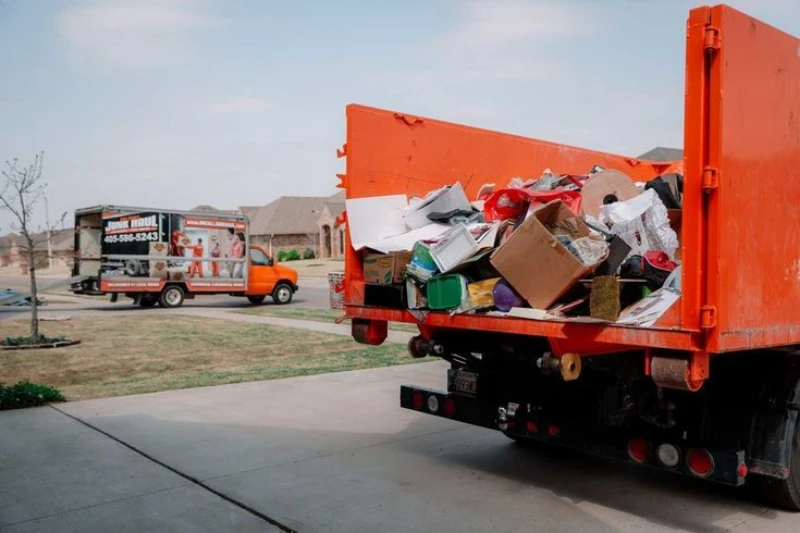 Orange garbage truck overflowing with cardboard and household waste on a suburban street; a moving truck and homes appear in the background.