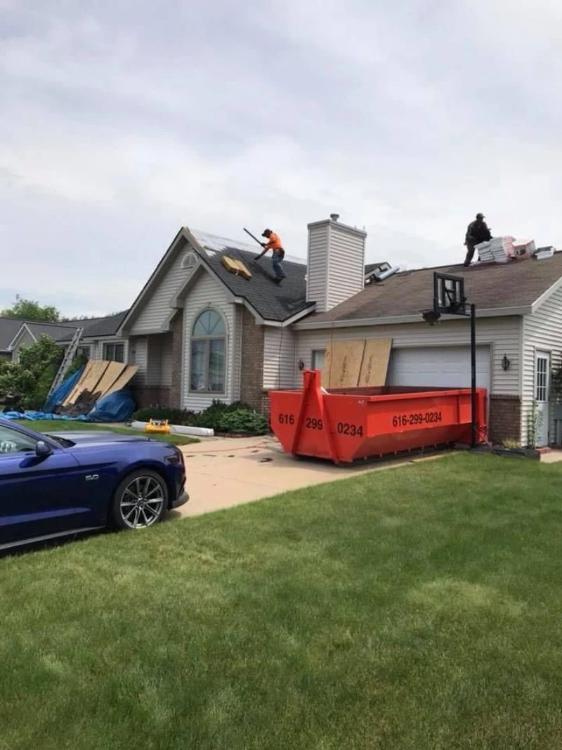 Workers repair a suburban house roof beside a red construction dumpster, scattered lumber, and a blue car under an overcast sky.