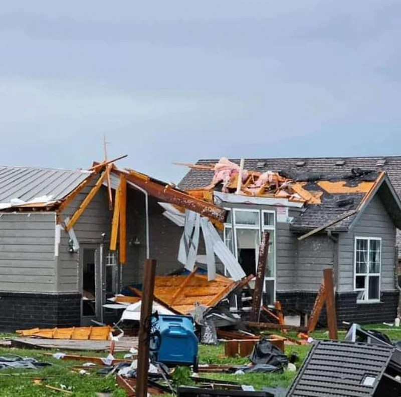 Storm-damaged gray house with a collapsed roof, broken windows, exposed rooms, and debris scattered across the lawn under a cloudy sky.