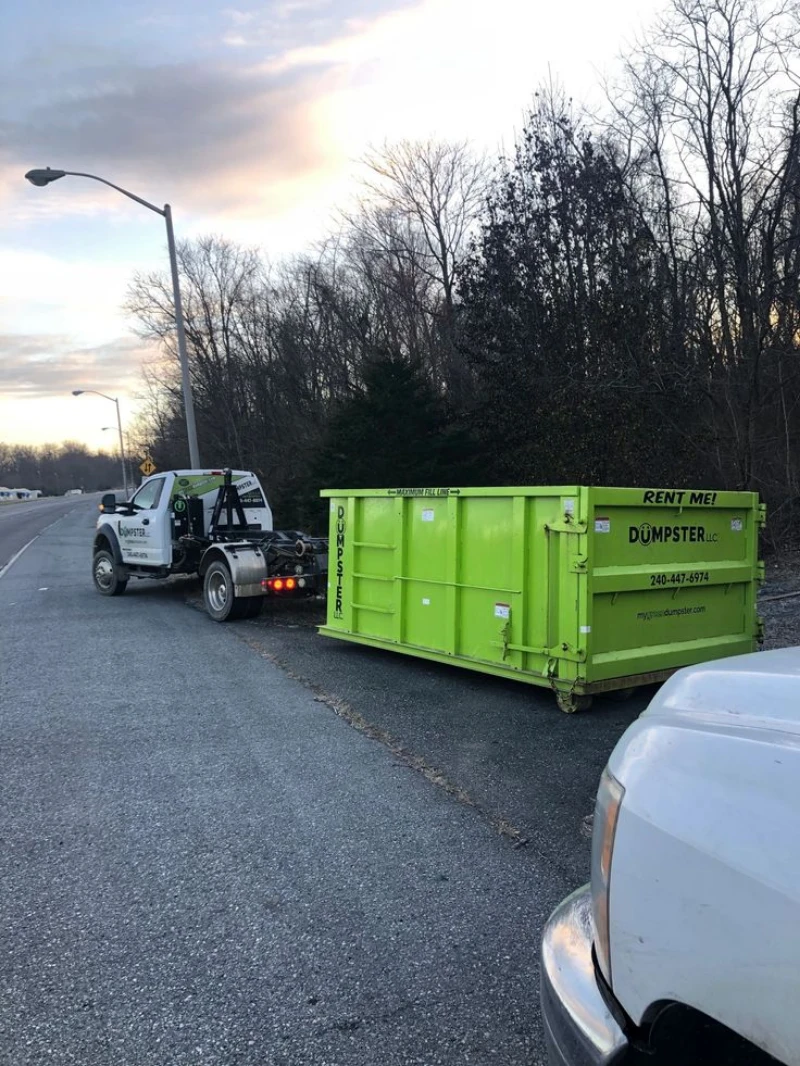 A tow truck hauls a bright green RENT ME dumpster along a tree-lined roadside at dusk, beneath a cloudy sky, creating a quiet mood.