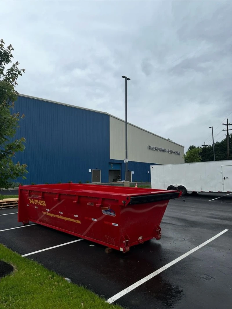 Bright red commercial dumpster sits in a wet, striped parking lot beside a blue-and-gray warehouse, with trees under a gloomy sky.