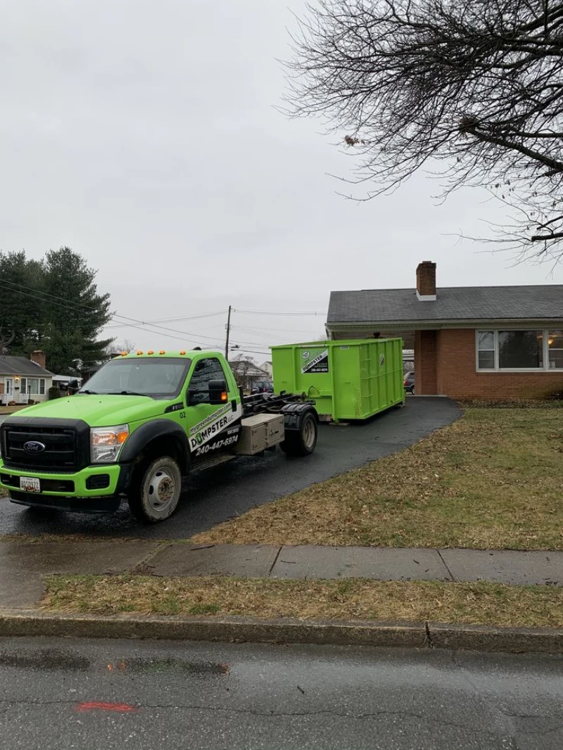 Green dumpster truck parked in a suburban driveway beside a brick house on a rainy, overcast day; wet street and bare trees create a somber mood.