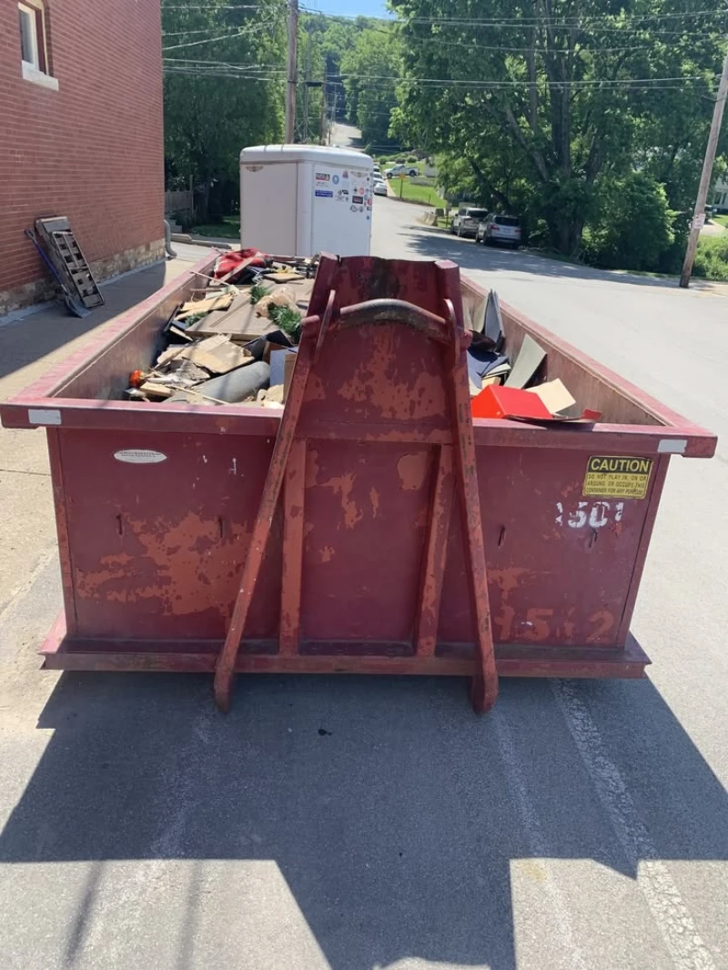 A rusty red construction dumpster filled with cardboard and debris sits on a sunny street beside brick buildings, trees, and utility lines.