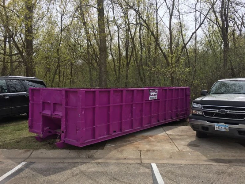 Large purple construction dumpster parked between two SUVs in a wooded parking lot, with budding trees and a calm springtime atmosphere.