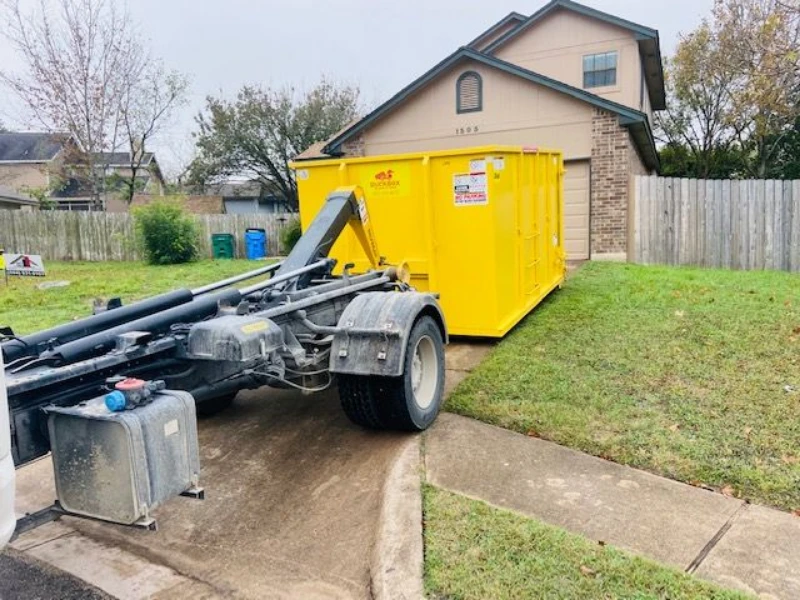 Roll-off truck positions a bright yellow dumpster beside a suburban home, driveway, lawn, and wooden fence under an overcast sky.