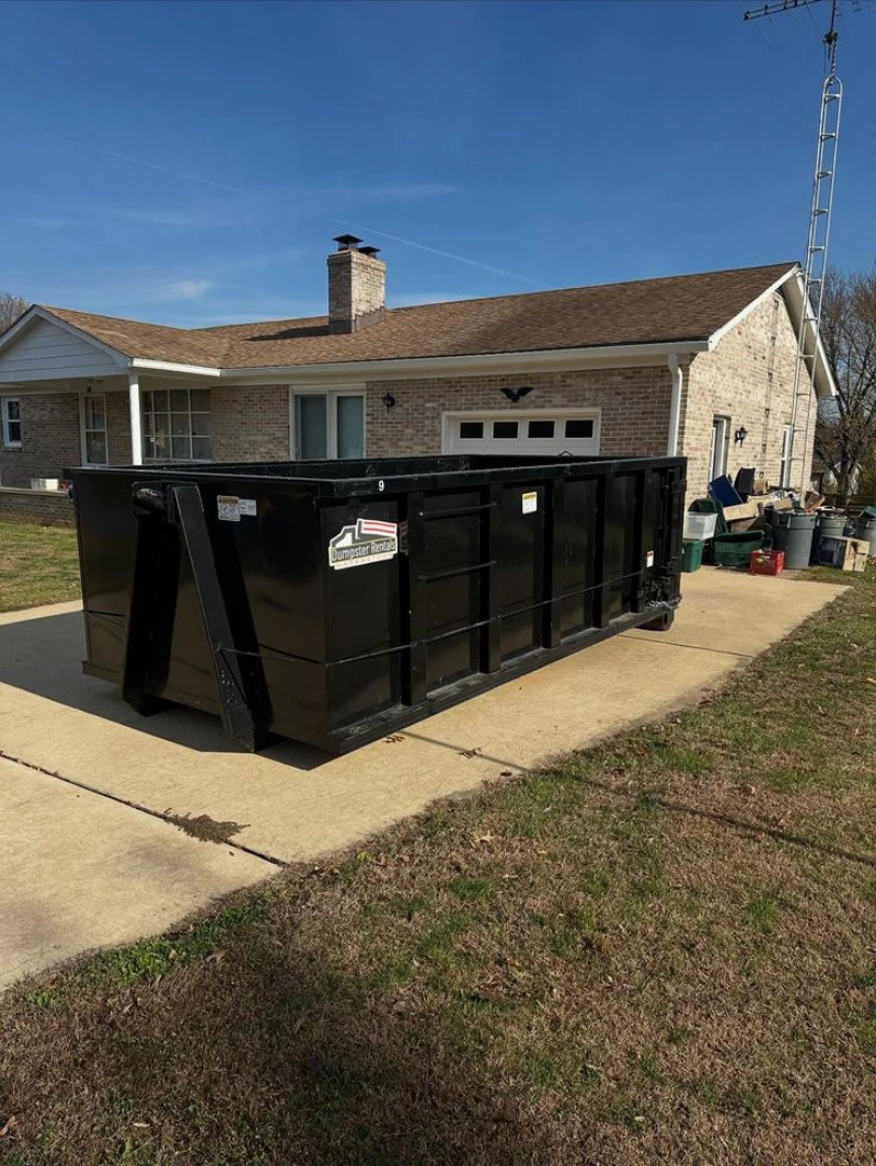 Large black commercial dumpster sits in a sunny residential driveway, with a brick house, lawn, recycling bins, and clear blue sky behind it.