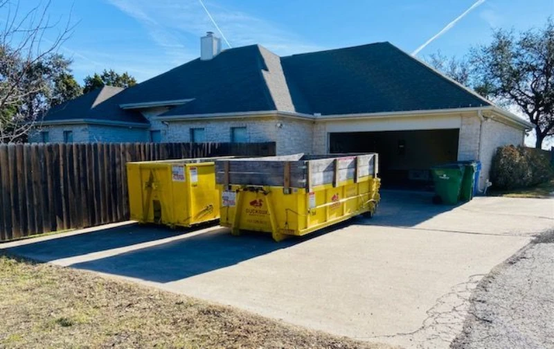 A suburban brick house with an open garage, wooden fence, and three large yellow construction dumpsters in the driveway under a bright blue sky.