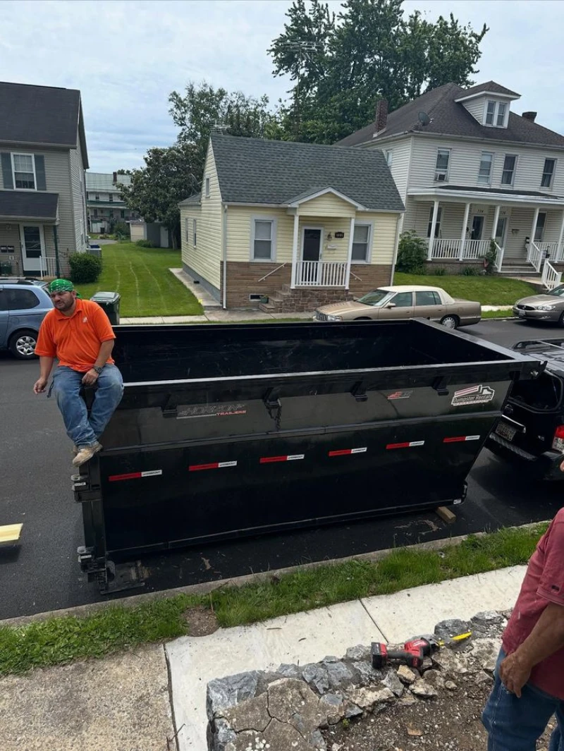 A worker in an orange shirt sits on a large black dumpster outside older homes, while another person and tools stand near a rocky sidewalk.