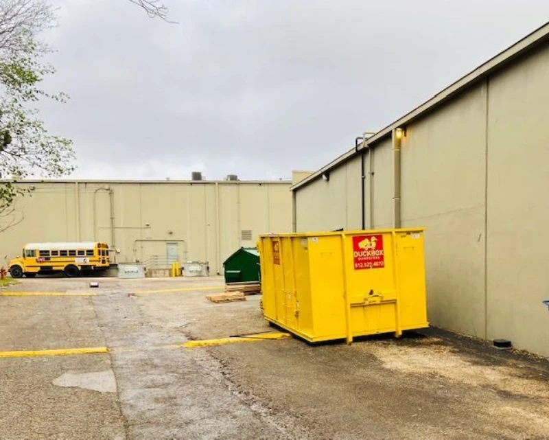 Industrial loading area beneath gray skies, featuring a bright yellow dumpster, parked school bus, warehouse walls, and damp pavement.