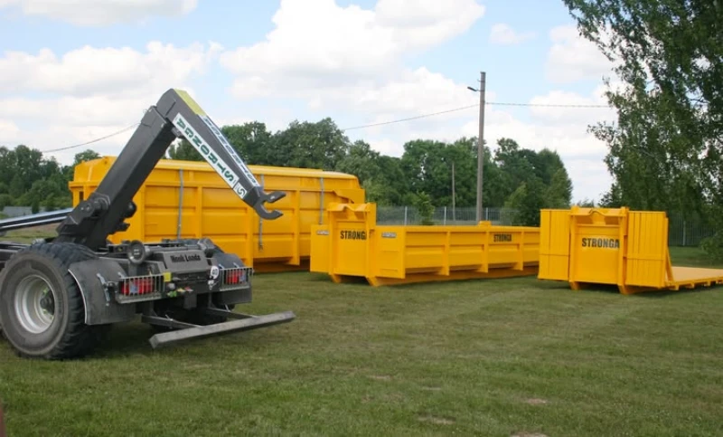 Black hook-lift truck parked beside bright yellow roll-off dumpsters on grass, with trees and blue sky creating a sunny outdoor industrial scene.