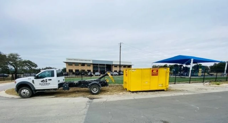 White flatbed truck parked beside a bright yellow dumpster outside a fenced facility with a blue canopy under an overcast sky.