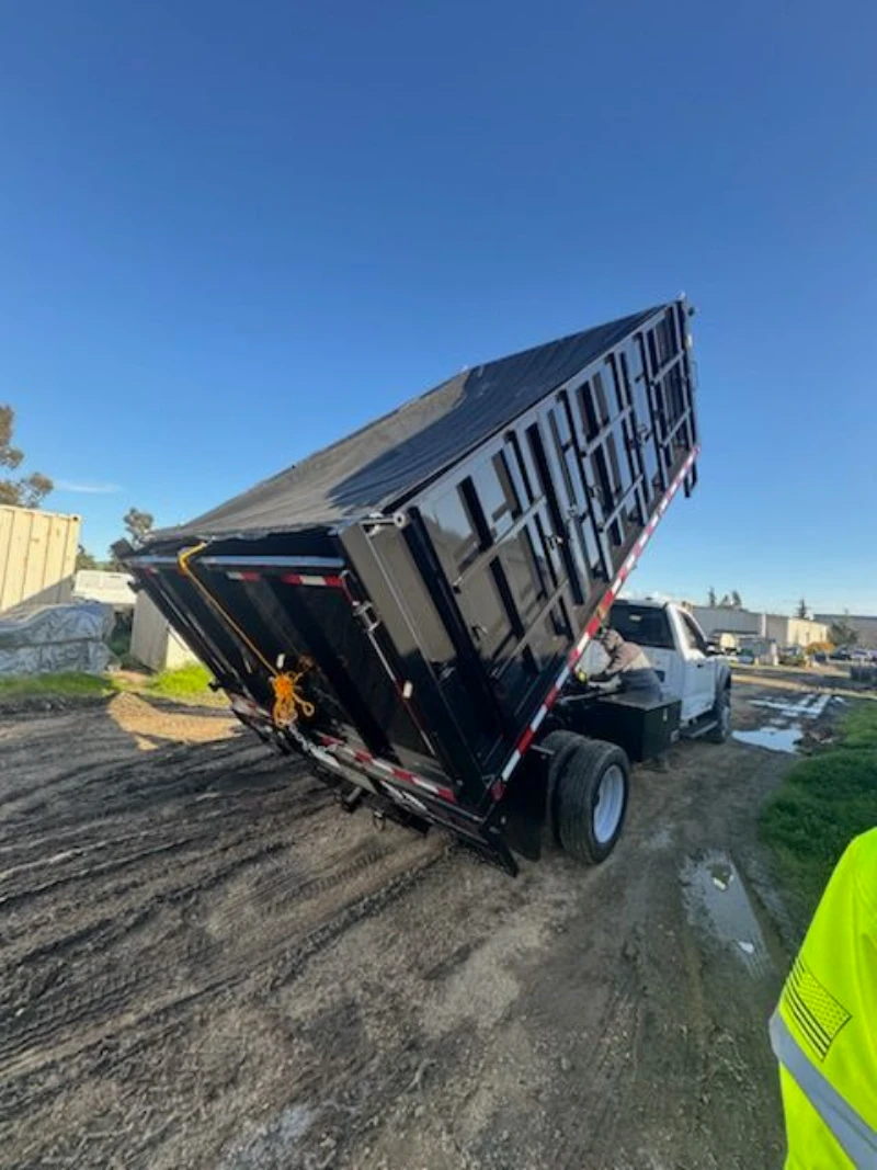 Raised black dump trailer attached to a white truck at a muddy worksite, with puddles, blue sky, and a worker in a neon safety vest nearby.