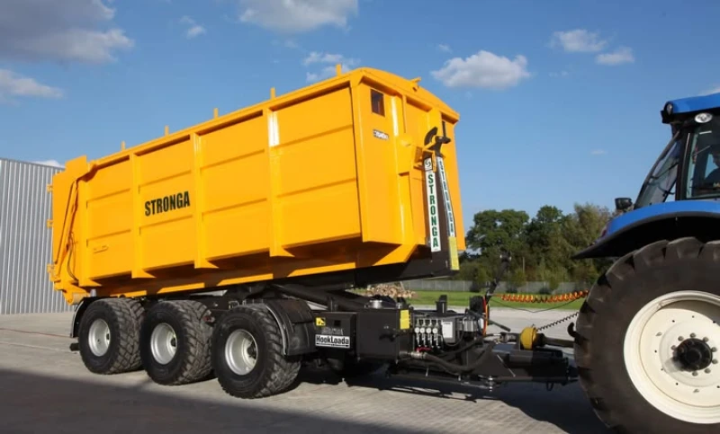 Bright yellow STRONGA agricultural trailer hitched to a blue tractor, shown on pavement beneath a sunny blue sky; rugged farm equipment.