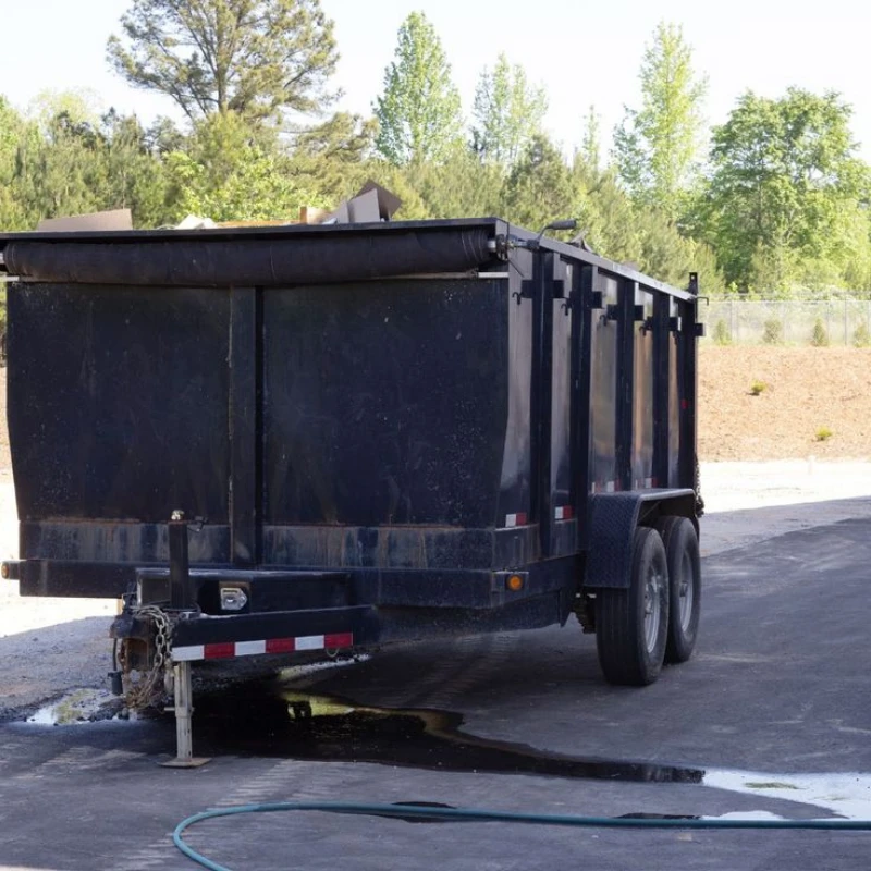 Large black utility dump trailer parked outdoors on pavement, with dual wheels, hitch, water hose, trees, and a quiet industrial setting.